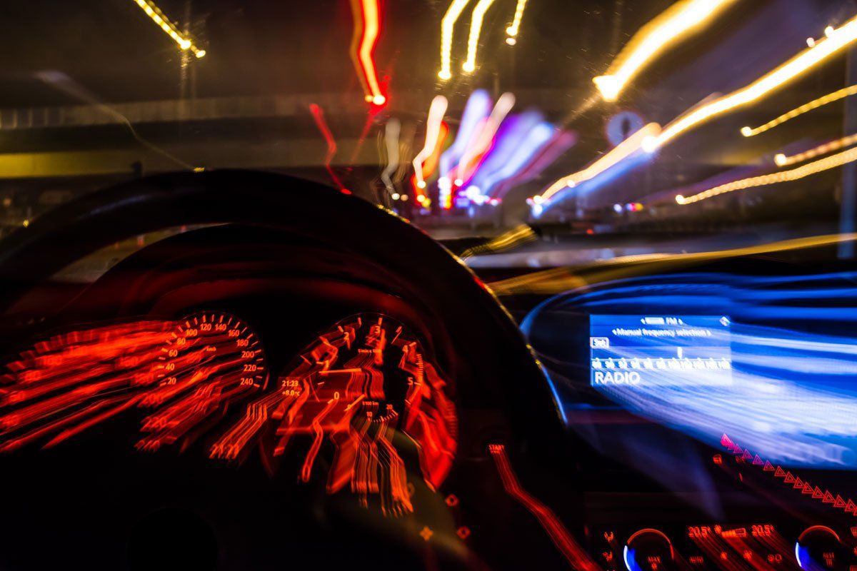 A driver's view from inside a car at night, showing motion-blurred street lights and illuminated dashboard controls.