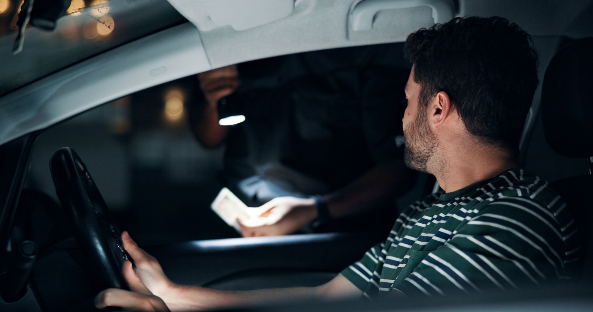 At night, a person in a striped shirt sits in a car while an officer shines a flashlight and inspects their ID.