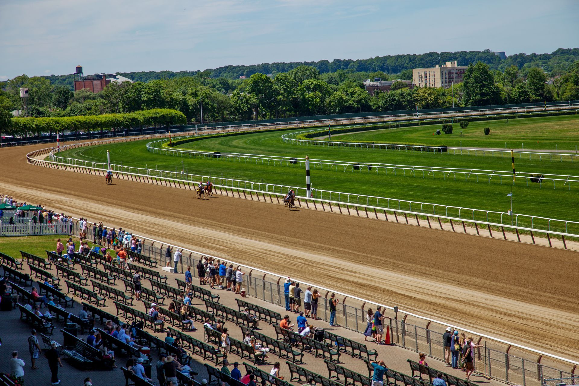 Racehorses on a dirt track, spectators in the stands, green grass field, trees, and buildings in the background.