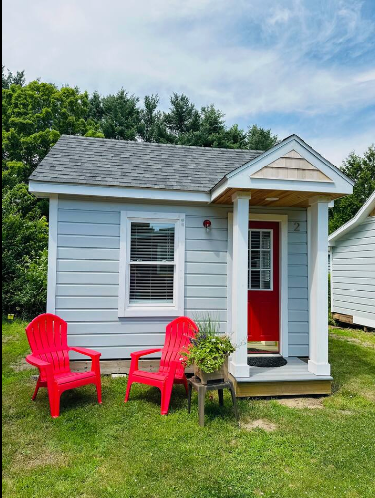Small blue cabin with red door and chairs on a grassy lawn.