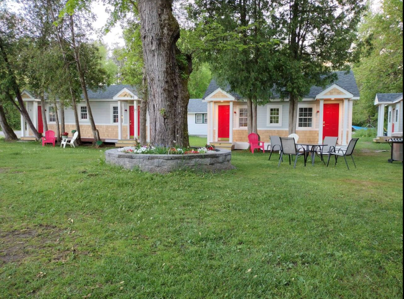 Small cabins with red doors, light blue walls, and a tree in front, with chairs on the grass.