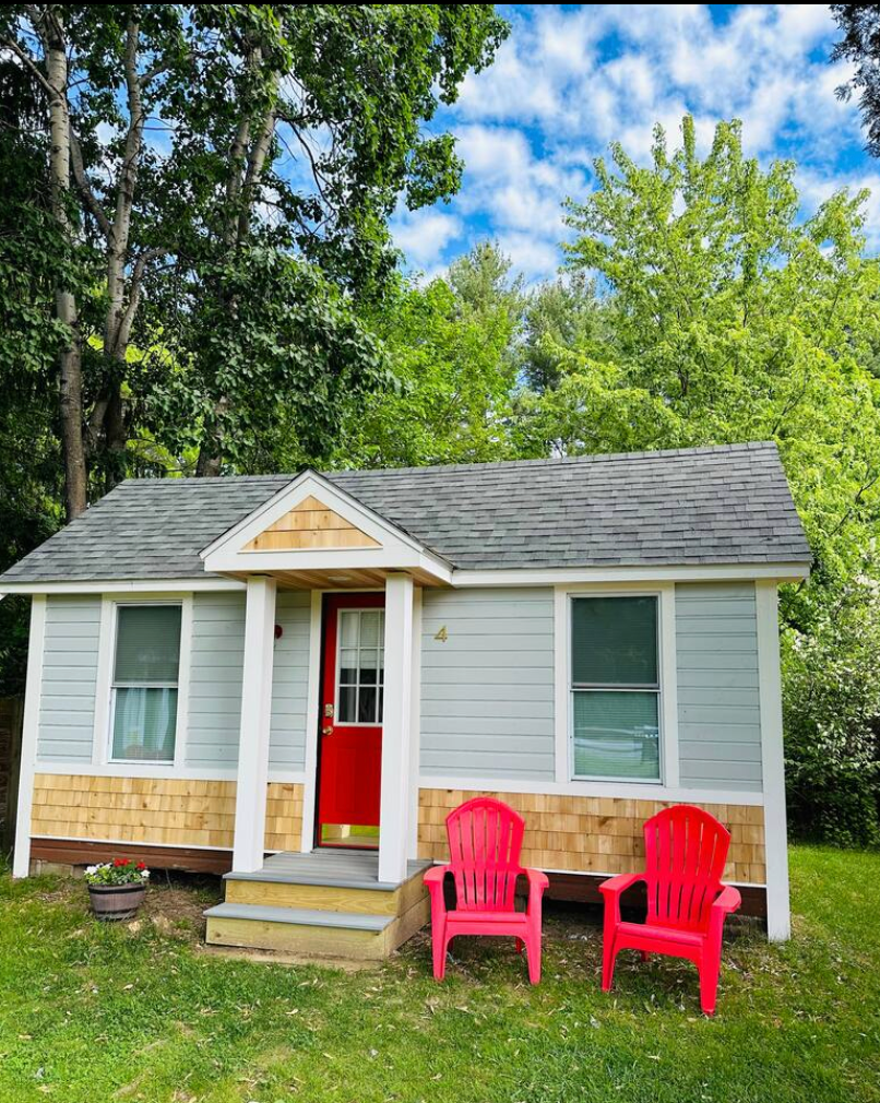 Small gray cabin with red door, cedar accents, two red chairs on lawn.