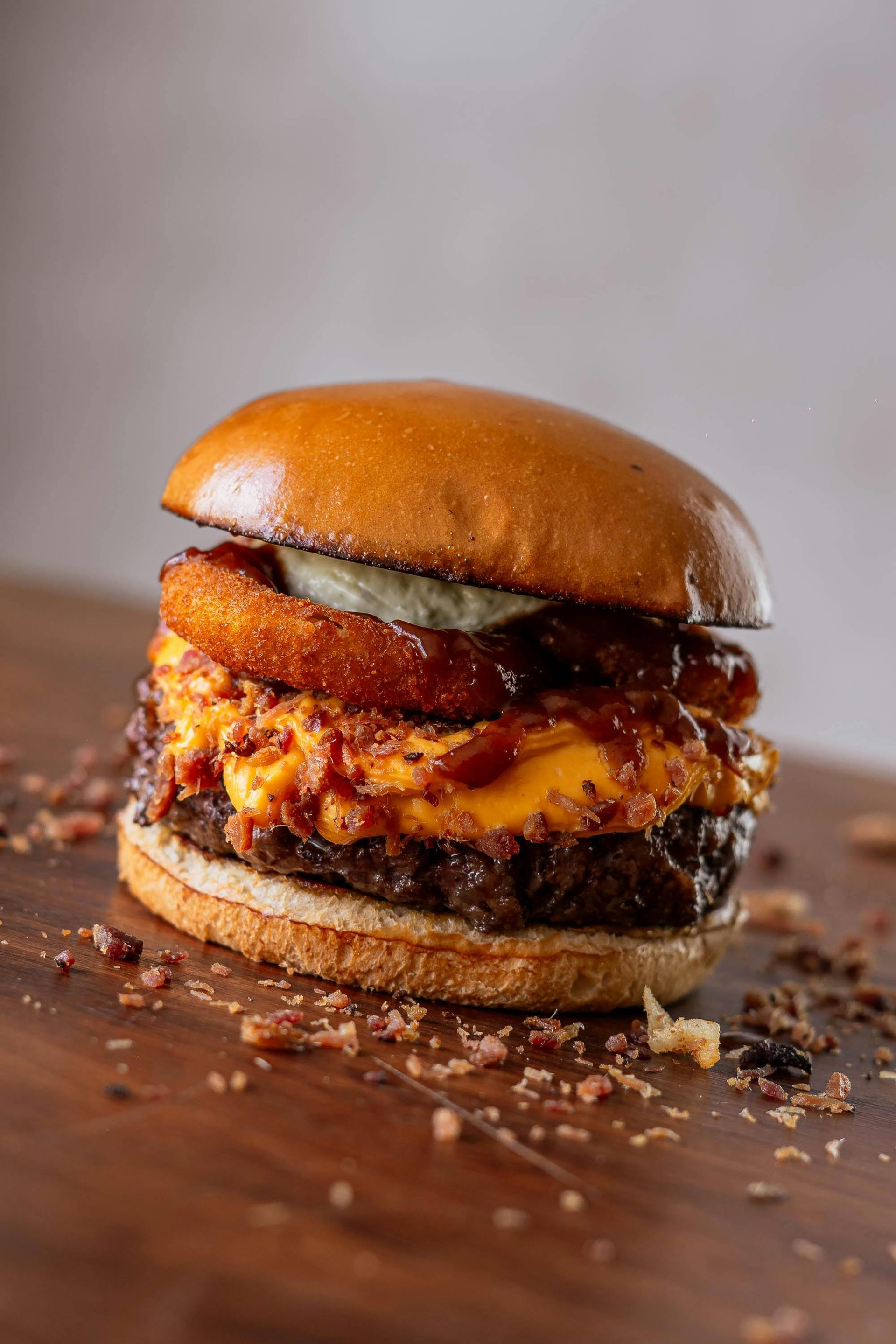Burger on a wooden surface with a top bun, onion ring, cheese, and a patty.