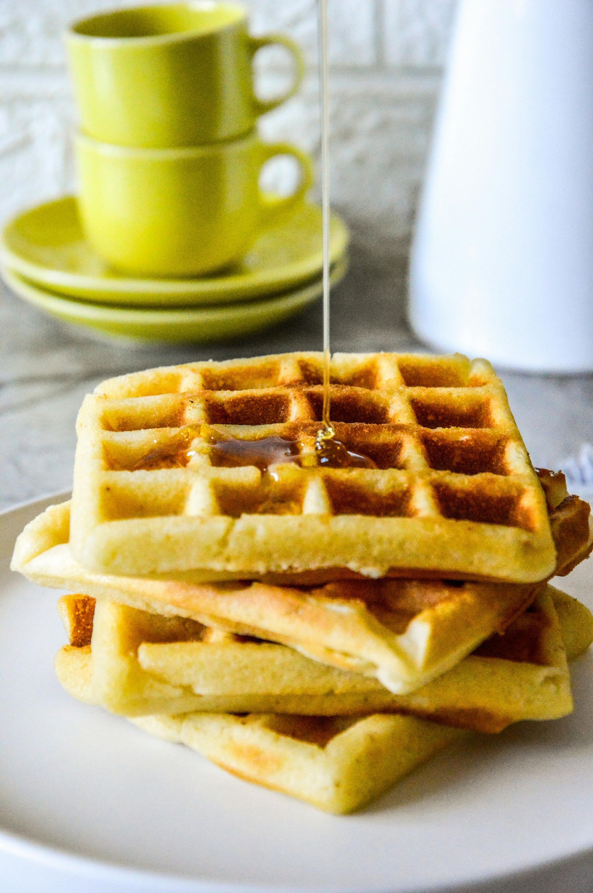Stack of golden waffles with syrup being poured on top, next to yellow cups and plates.