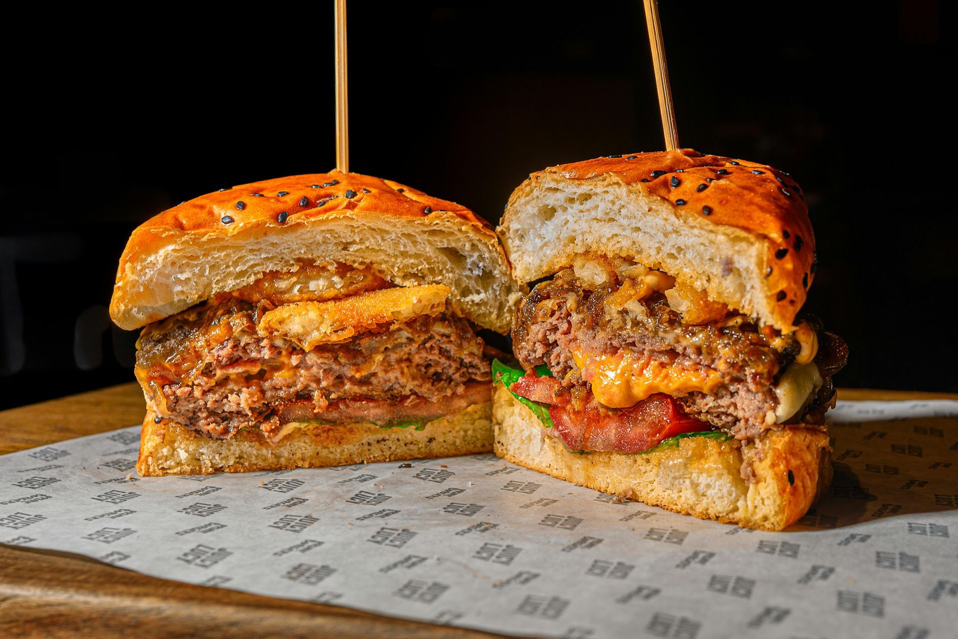 Burger cut in half, showing meat, cheese, and toppings on a sesame seed bun, resting on printed paper.