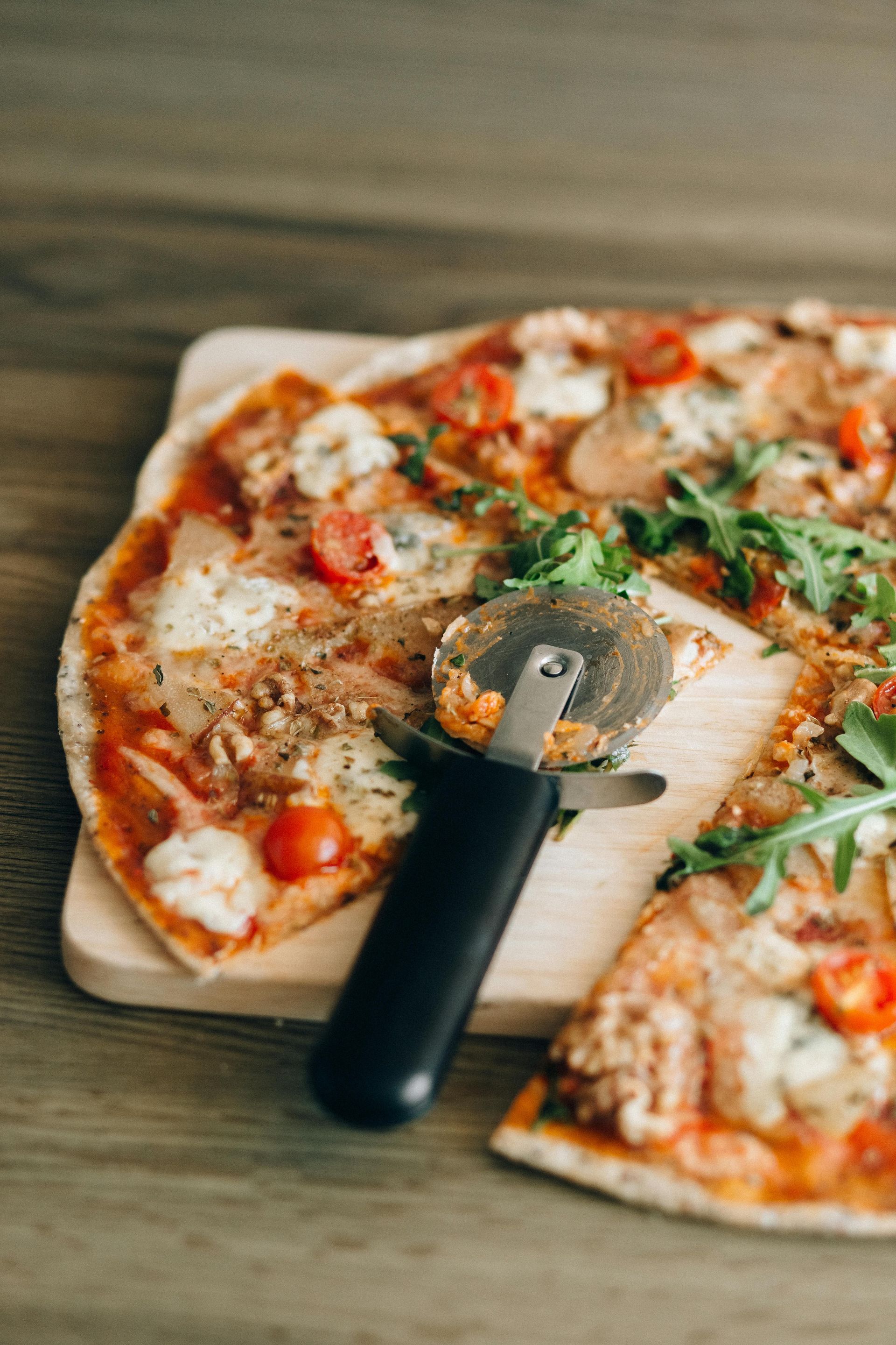Pizza on wooden board with a pizza cutter partially slicing a piece.