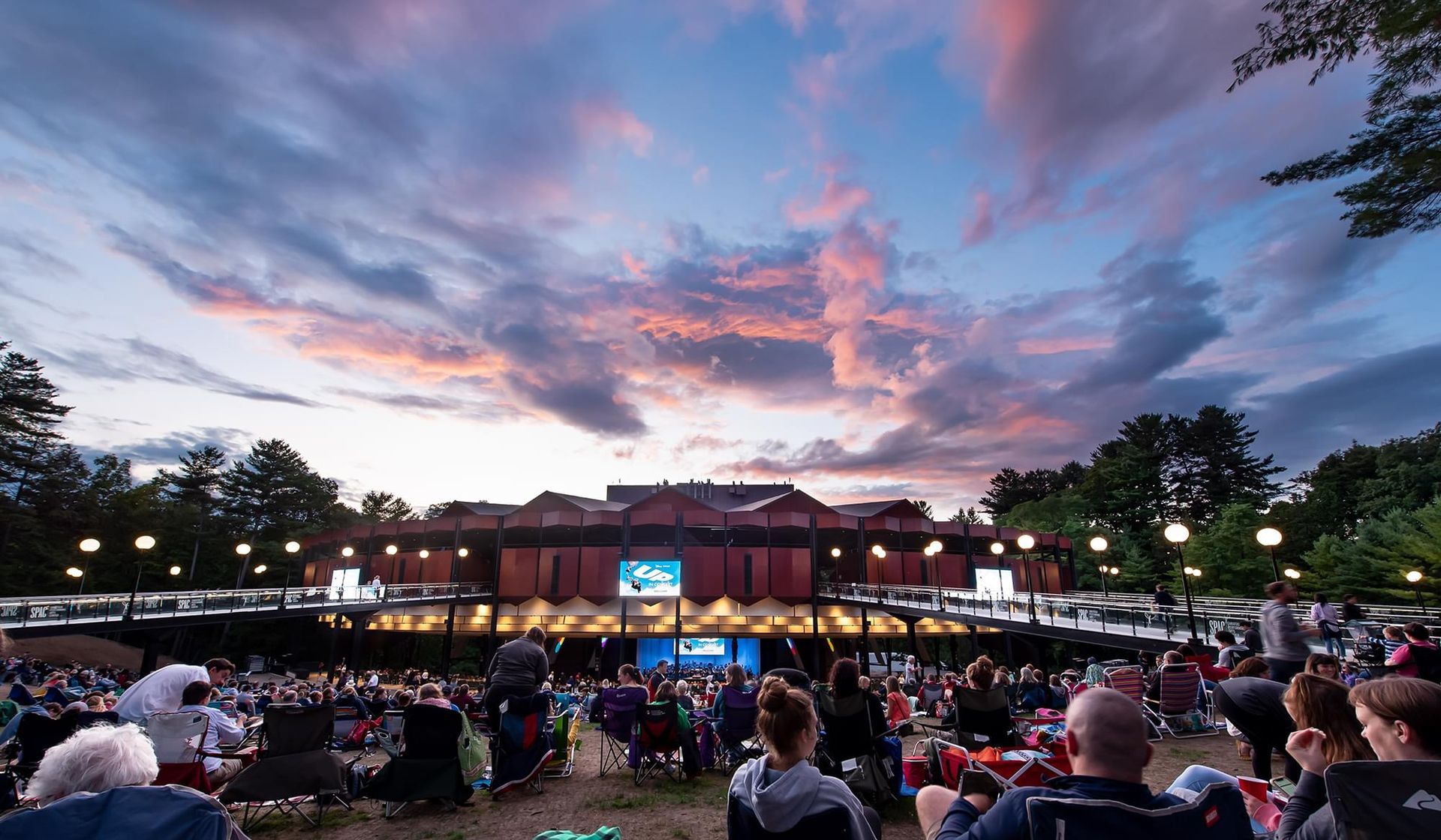 Outdoor theater with audience seated, under a sunset sky. Building is in the background.