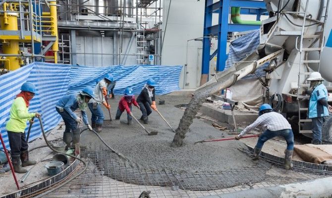Construction workers spreading wet concrete at a building site with machinery and blue tarp background