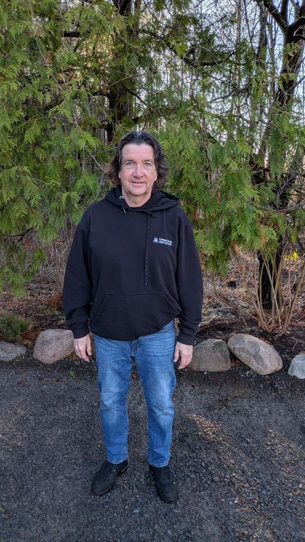 Person standing outdoors in a black hoodie and blue jeans in front of trees and rocks