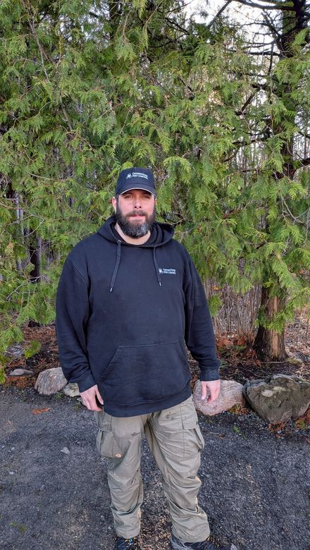 Person standing outdoors in a black hoodie and cap in front of green foliage.