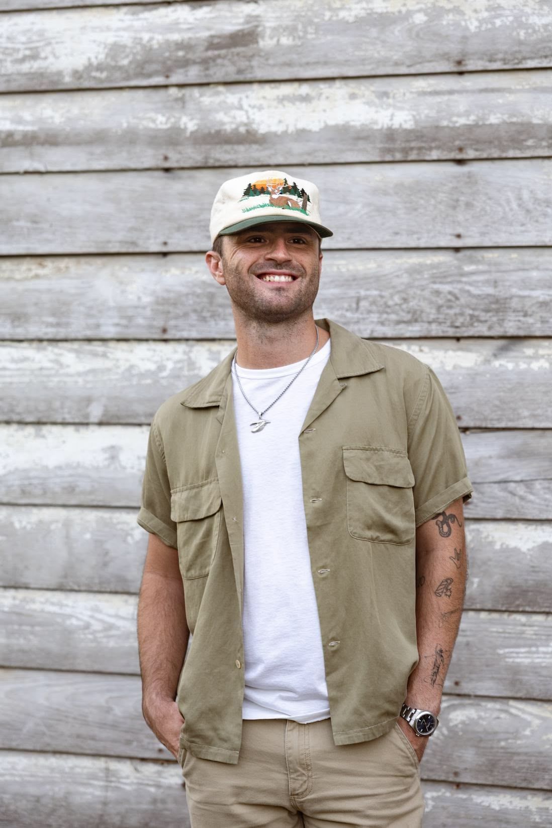 A smiling person wearing a graphic cap, olive green short-sleeved button-up shirt, and white tee against wood paneling.