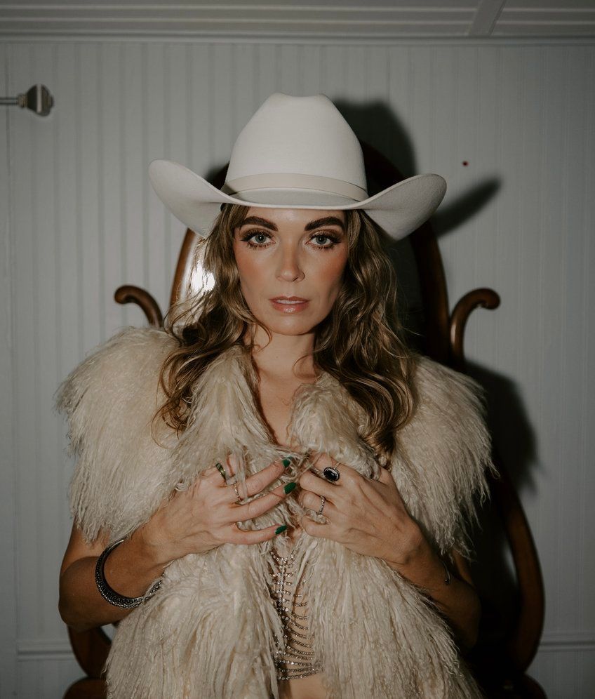 A person wearing a white cowboy hat and a tan fringe vest, looking at the camera against a plain, dimly lit background.