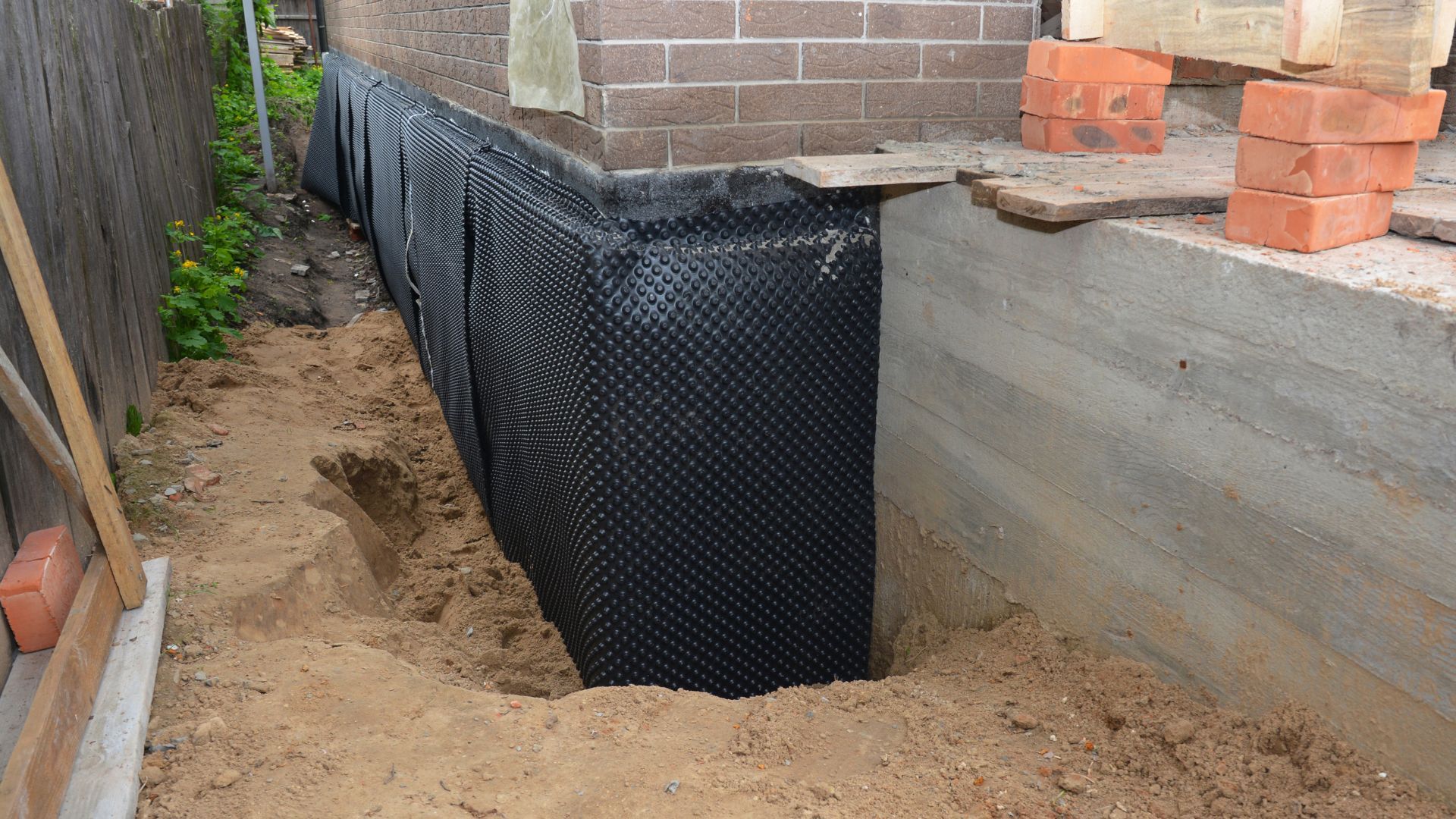Excavated foundation trench with black drainage membrane beside a concrete wall and bricks on top.