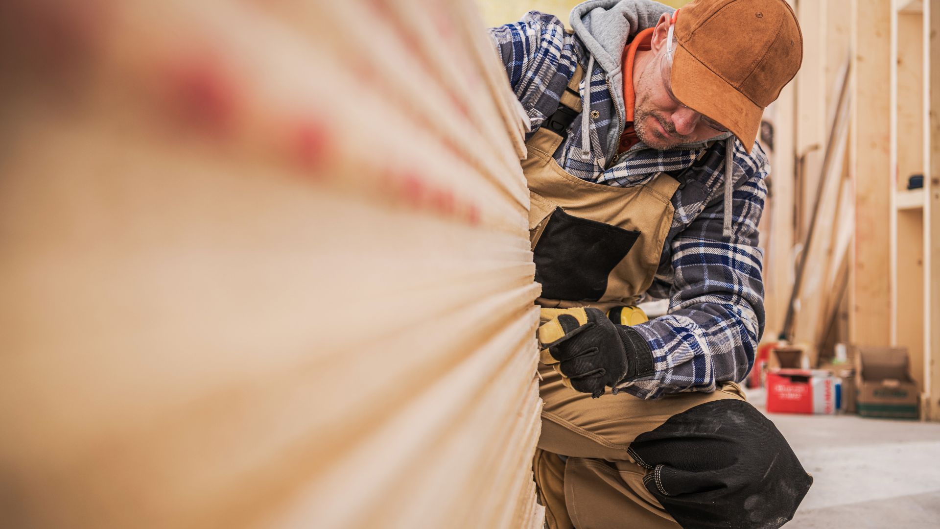 Construction worker fastening wood panel indoors with drill, wearing plaid shirt, orange hard hat, and gloves