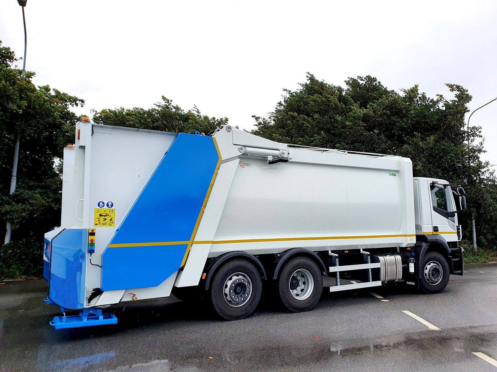 Camion della spazzatura bianco con una striscia blu parcheggiato su una strada vicino agli alberi.