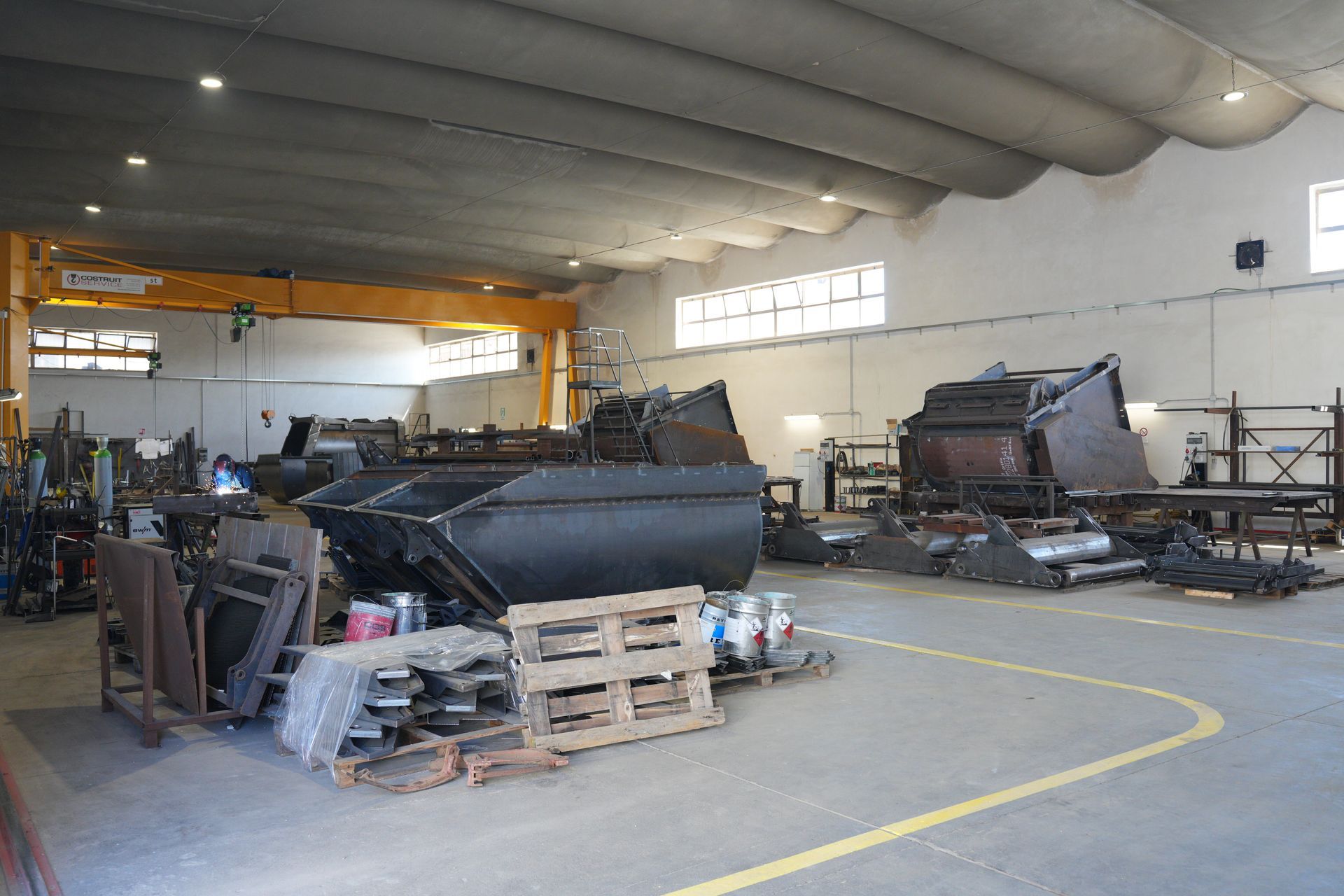 Officina industriale con grandi componenti metallici, pallet e macchinari sotto un alto soffitto curvo.