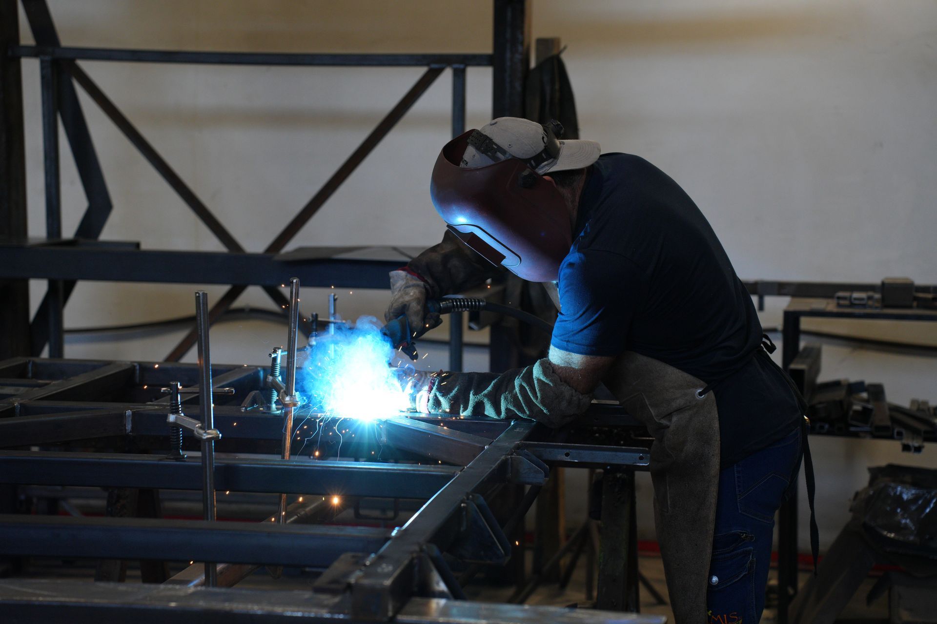 Un saldatore con indumenti protettivi sta saldando una struttura metallica; scintille blu brillanti si sprigionano nell'officina.