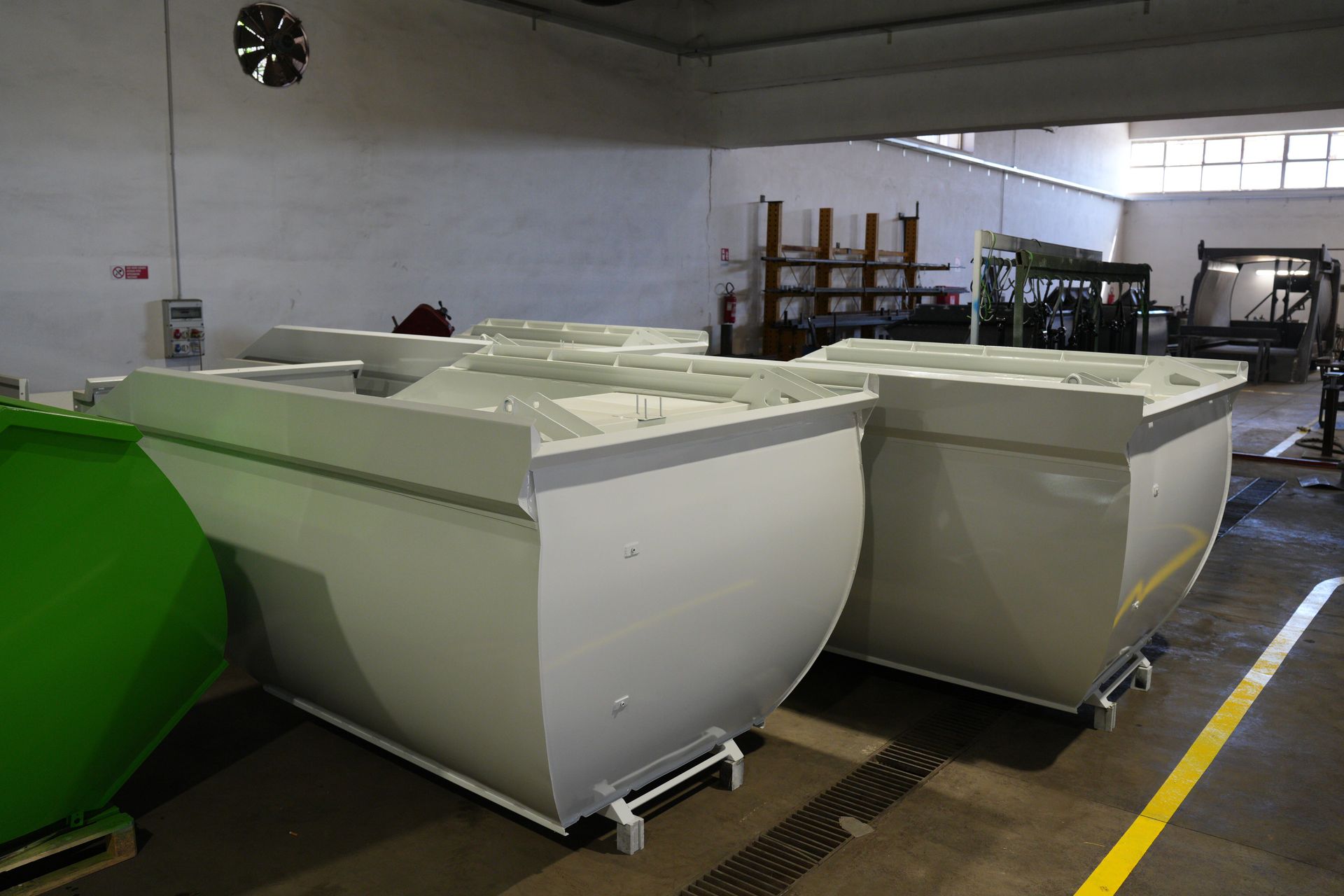 Grandi cassonetti industriali bianchi in un magazzino, con una cisterna verde sulla sinistra e macchinari sullo sfondo.