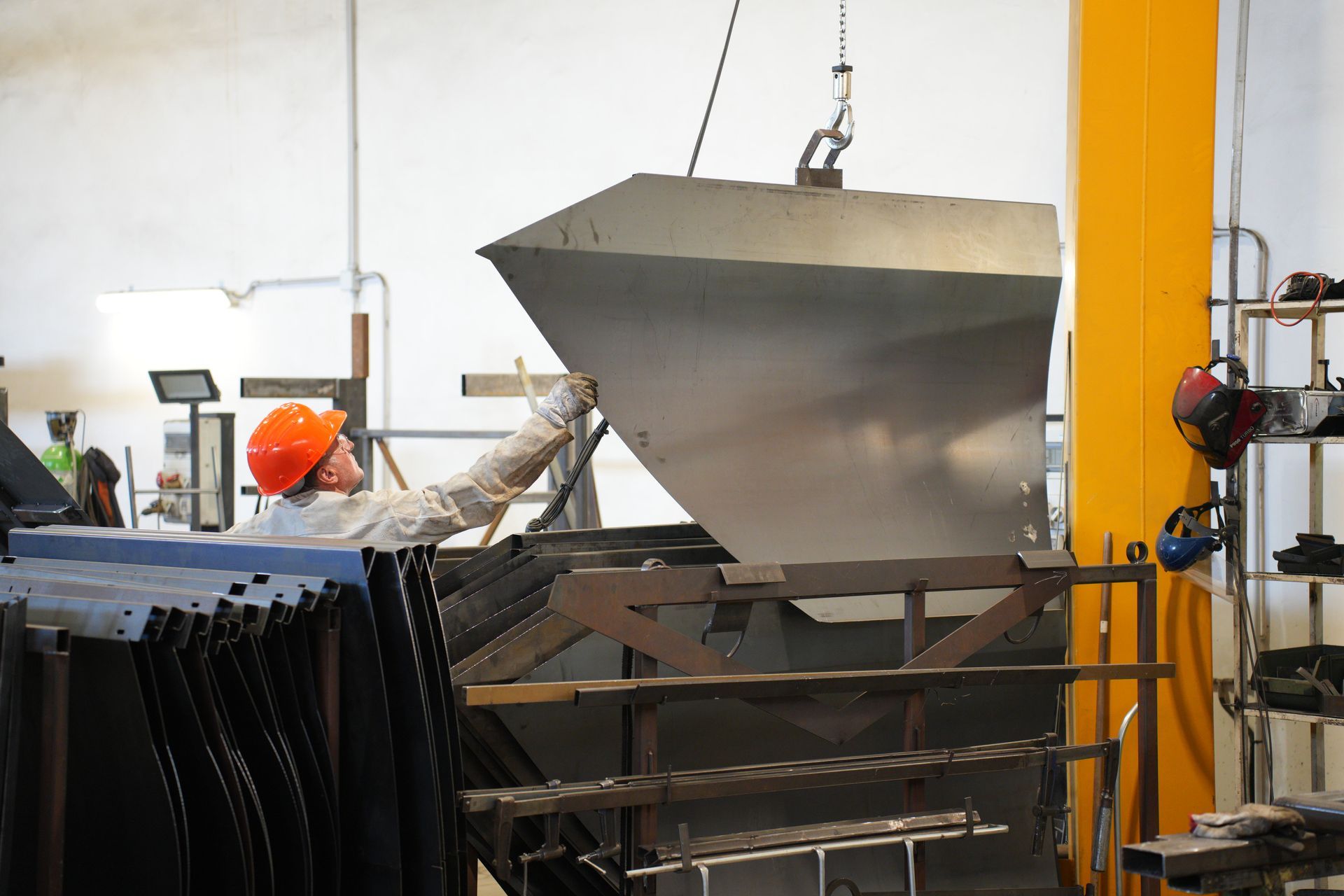 Una tramoggia industriale viene sollevata in un'officina da una gru accanto a travi di supporto gialle.