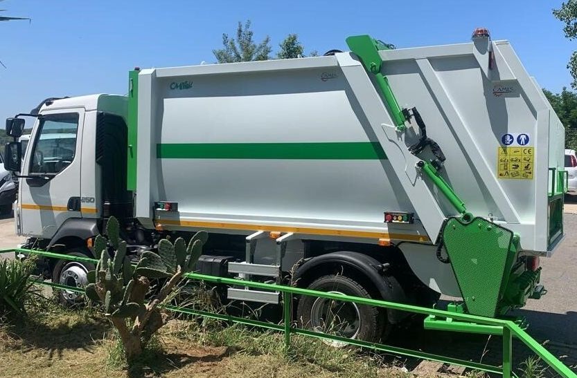 Un camion della spazzatura verde e bianco è parcheggiato all'aperto vicino a un albero frondoso e a una recinzione.