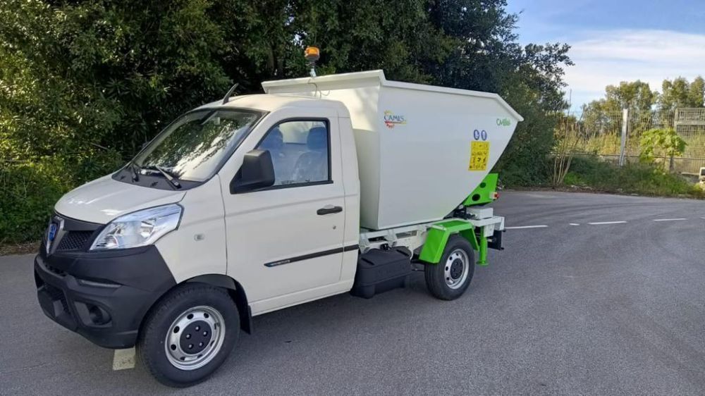 Un camioncino bianco con uno spargitore verde è parcheggiato a bordo strada vicino ad alcuni alberi.