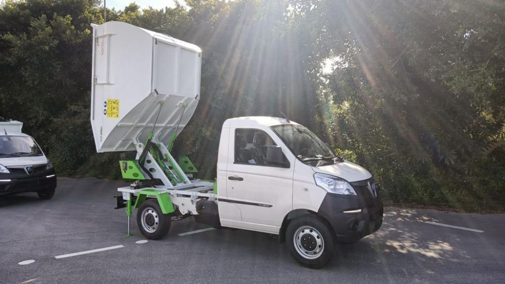 Un piccolo camioncino bianco con un pianale rialzato verde che trasporta un cassonetto bianco in un parcheggio soleggiato.