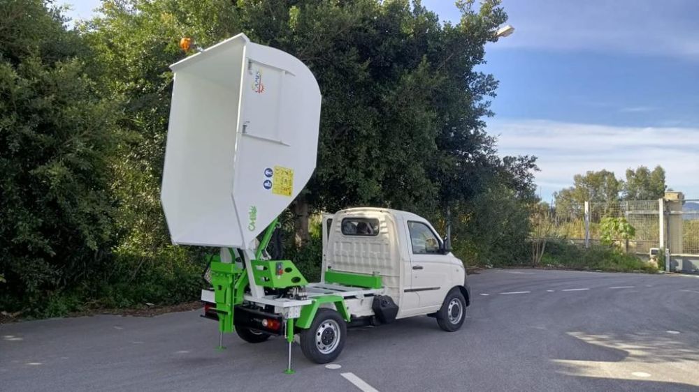 Piccolo camioncino bianco con pianale rialzato verde parcheggiato su una strada asfaltata accanto ad alberii.