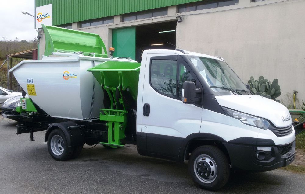 Camion della spazzatura bianco e verde parcheggiato accanto a un edificio con una tramoggia rialzata.