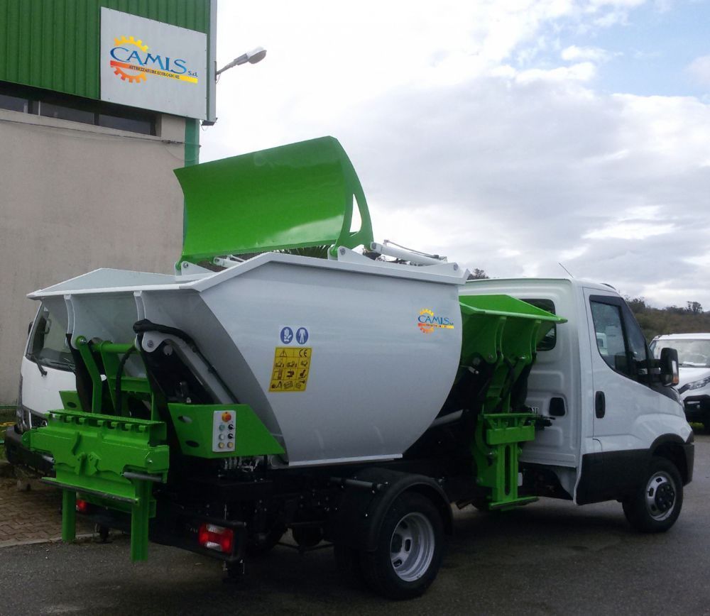 Camion bianco per la raccolta dei rifiuti con cassone verde e carrozzeria grigia parcheggiato all'aperto vicino a un edificio