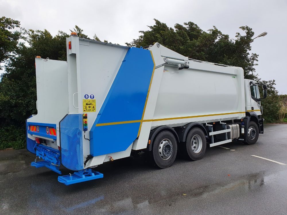 Camion della spazzatura bianco con pannello posteriore blu parcheggiato su una strada bagnata vicino agli alberi.