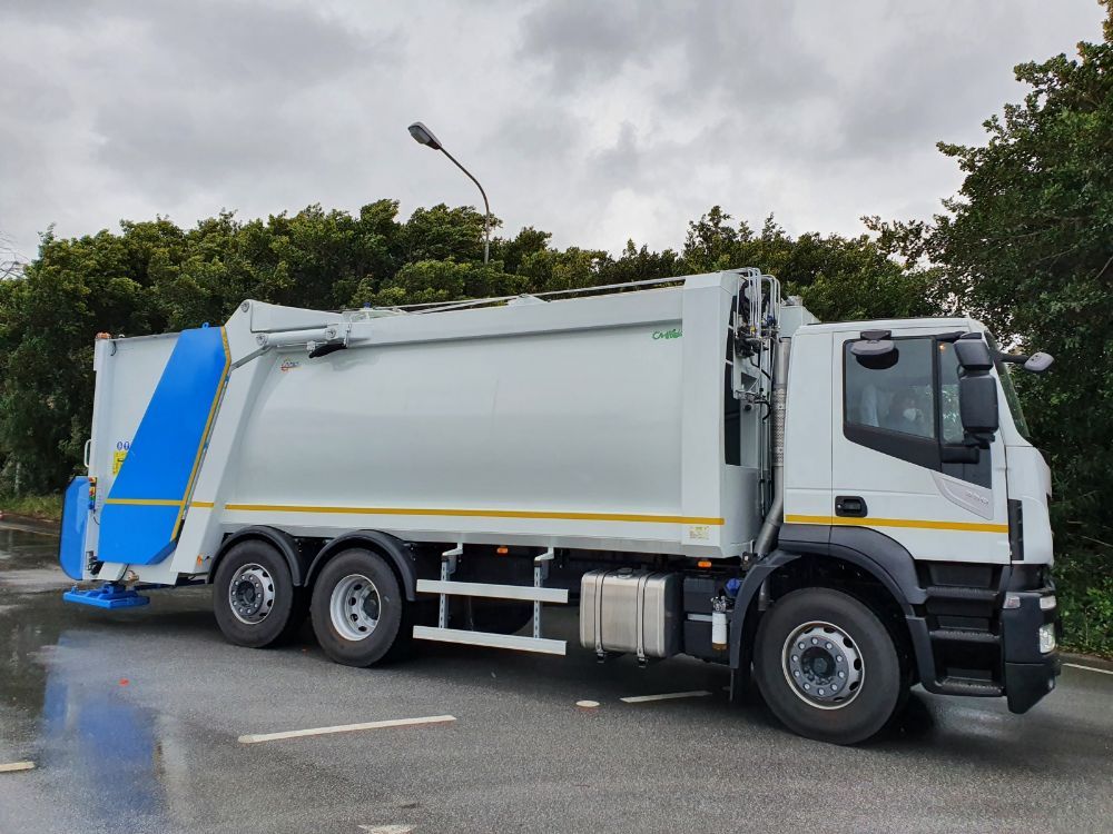 Camion della spazzatura bianco con telone posteriore blu parcheggiato su una strada bagnata sotto un cielo nuvoloso