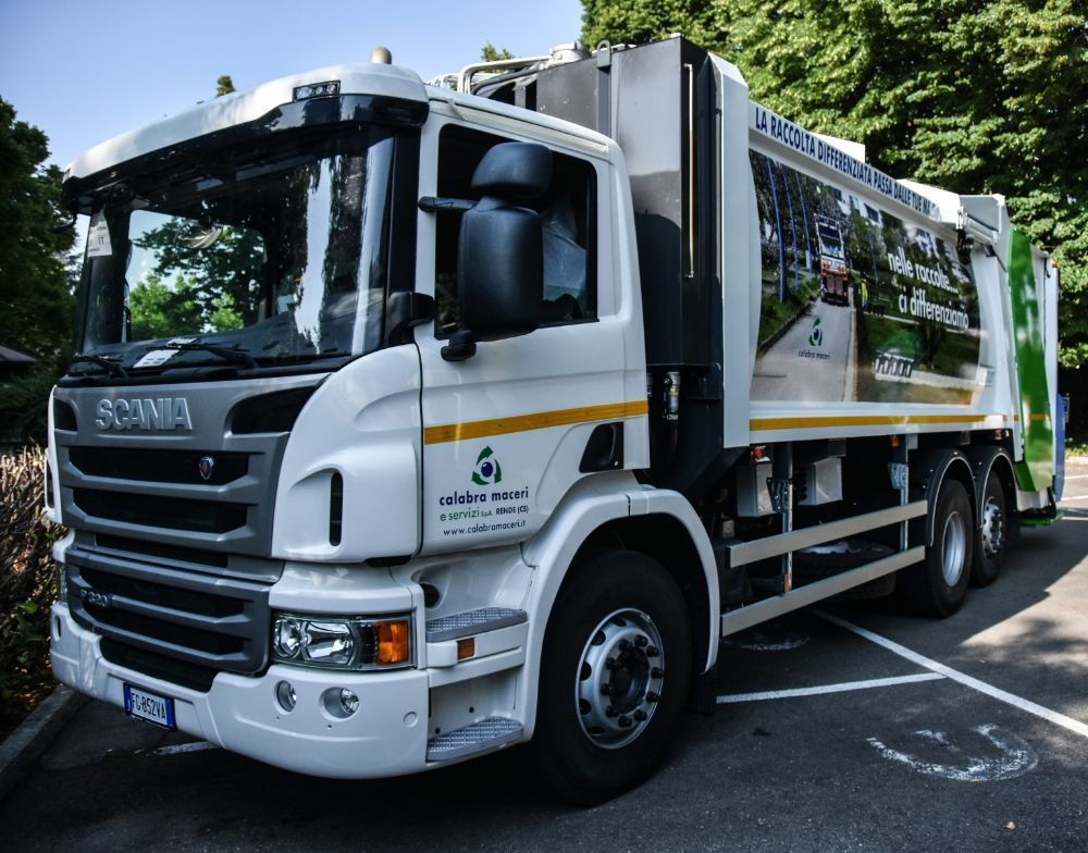 Un camion della spazzatura Scania bianco parcheggiato sul ciglio della strada, con alberi verdi sullo sfondo.