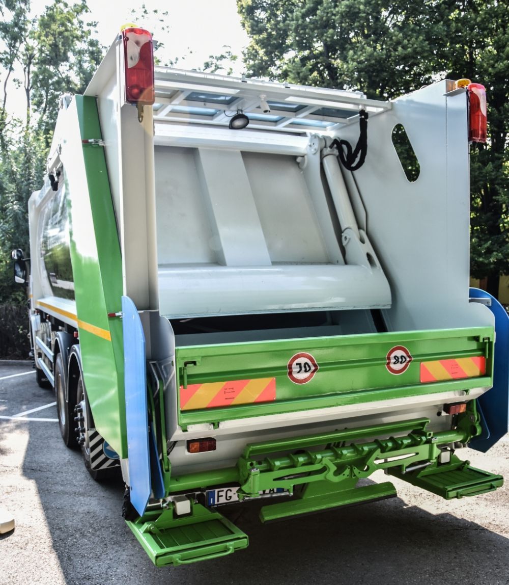 Vista posteriore di un camion della spazzatura verde e bianco con il cassone aperto su una strada alberata.