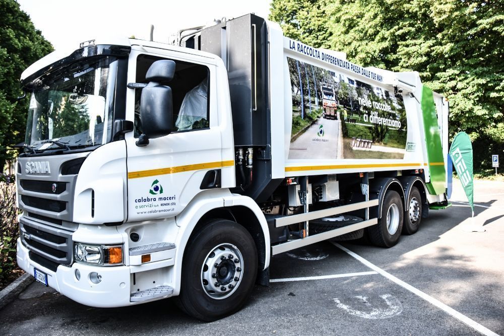 Camion della spazzatura bianco parcheggiato in strada, con grafiche sui pannelli laterali e bidoni verdi per i rifiuti sul retro.