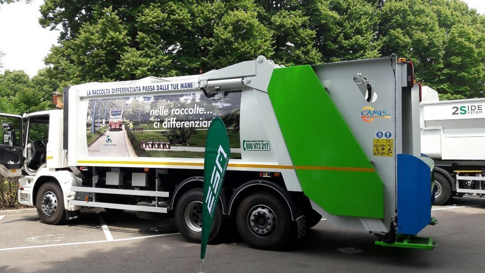 Camion della spazzatura verde e bianco parcheggiato all'aperto con un grande cassone posteriore e grafiche sui pannelli laterali.