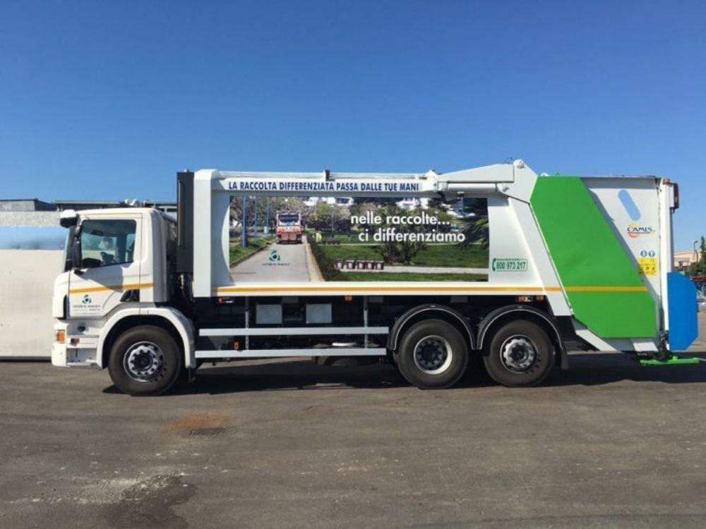 Camion della spazzatura bianco con pannello laterale verde parcheggiato sul marciapiede sotto un cielo azzurro e limpido.