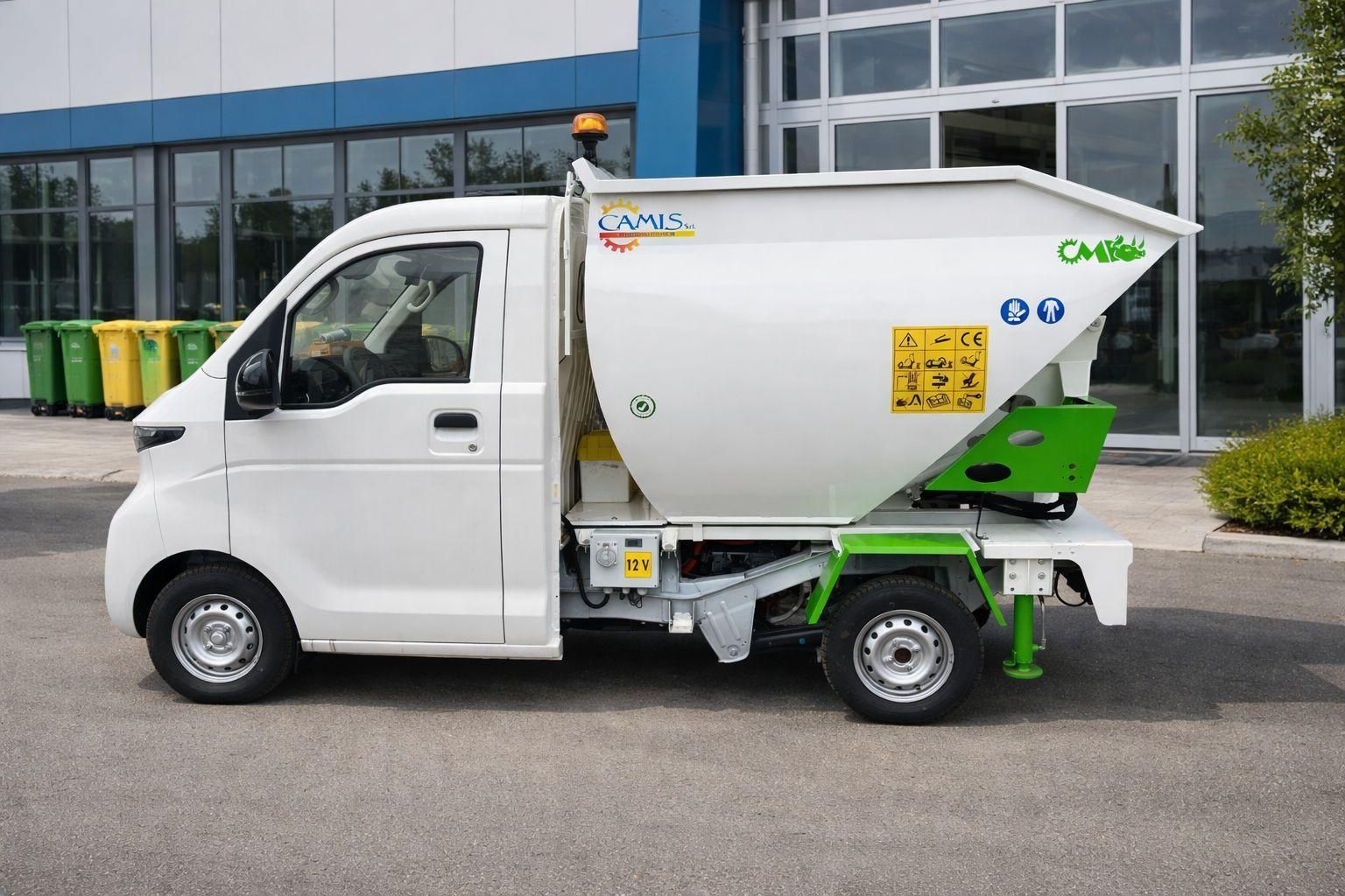 Un camioncino bianco compatto per la raccolta dei rifiuti è parcheggiato davanti a un edificio con finestre di vetro e cassonetti verdi nelle vicinanze.