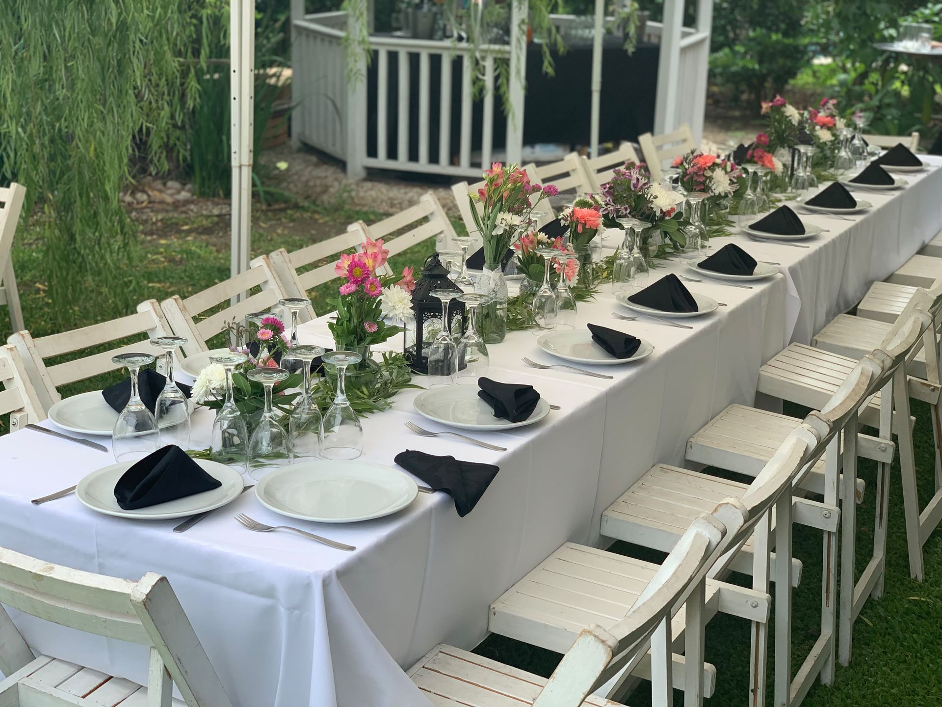 Larga mesa de banquete al aire libre con manteles blancos, servilletas negras, flores y sillas blancas en un jardín