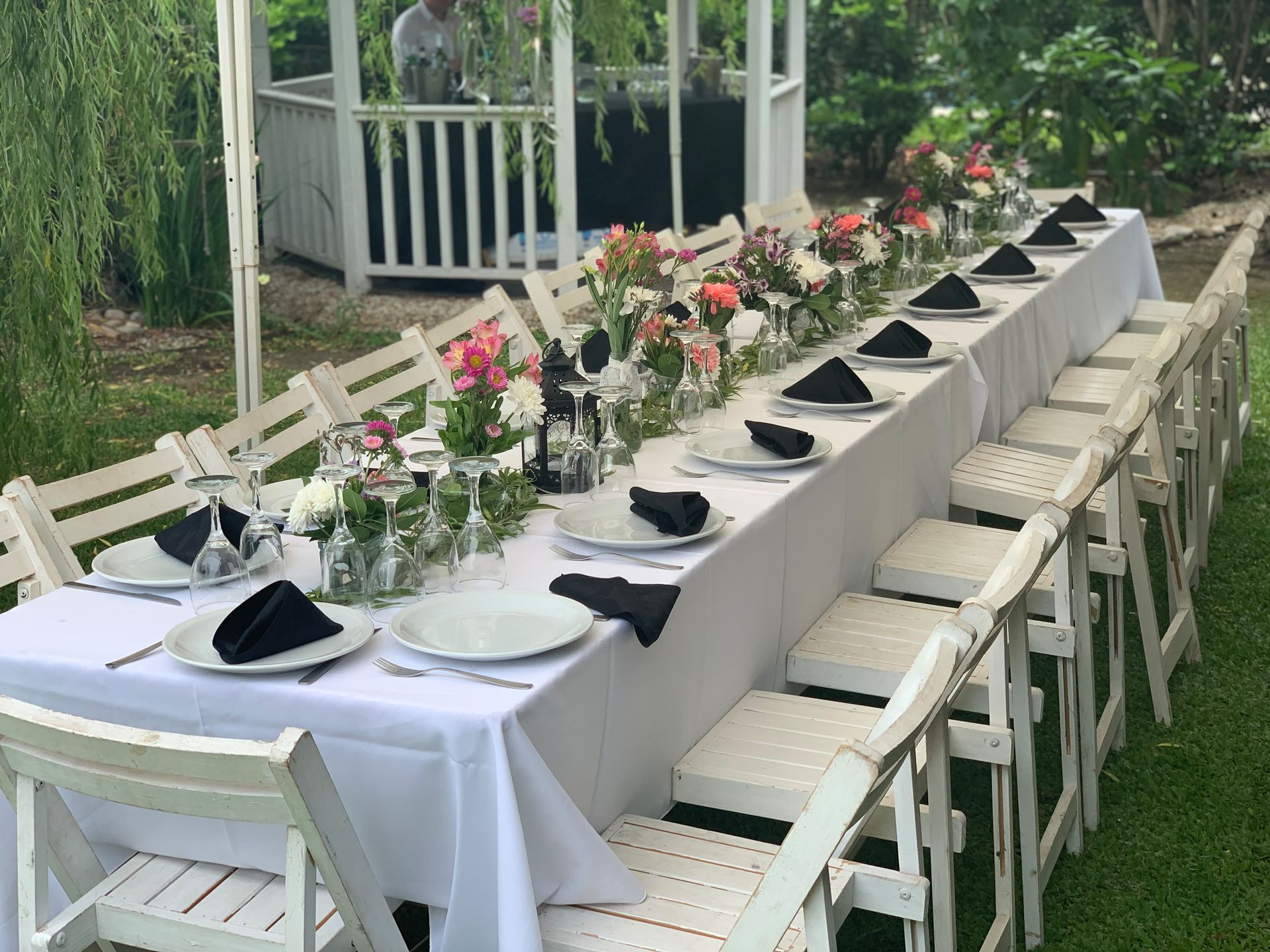 Mesa larga de comedor al aire libre con manteles blancos, servilletas negras y centros de mesa florales en una glorieta del jardín