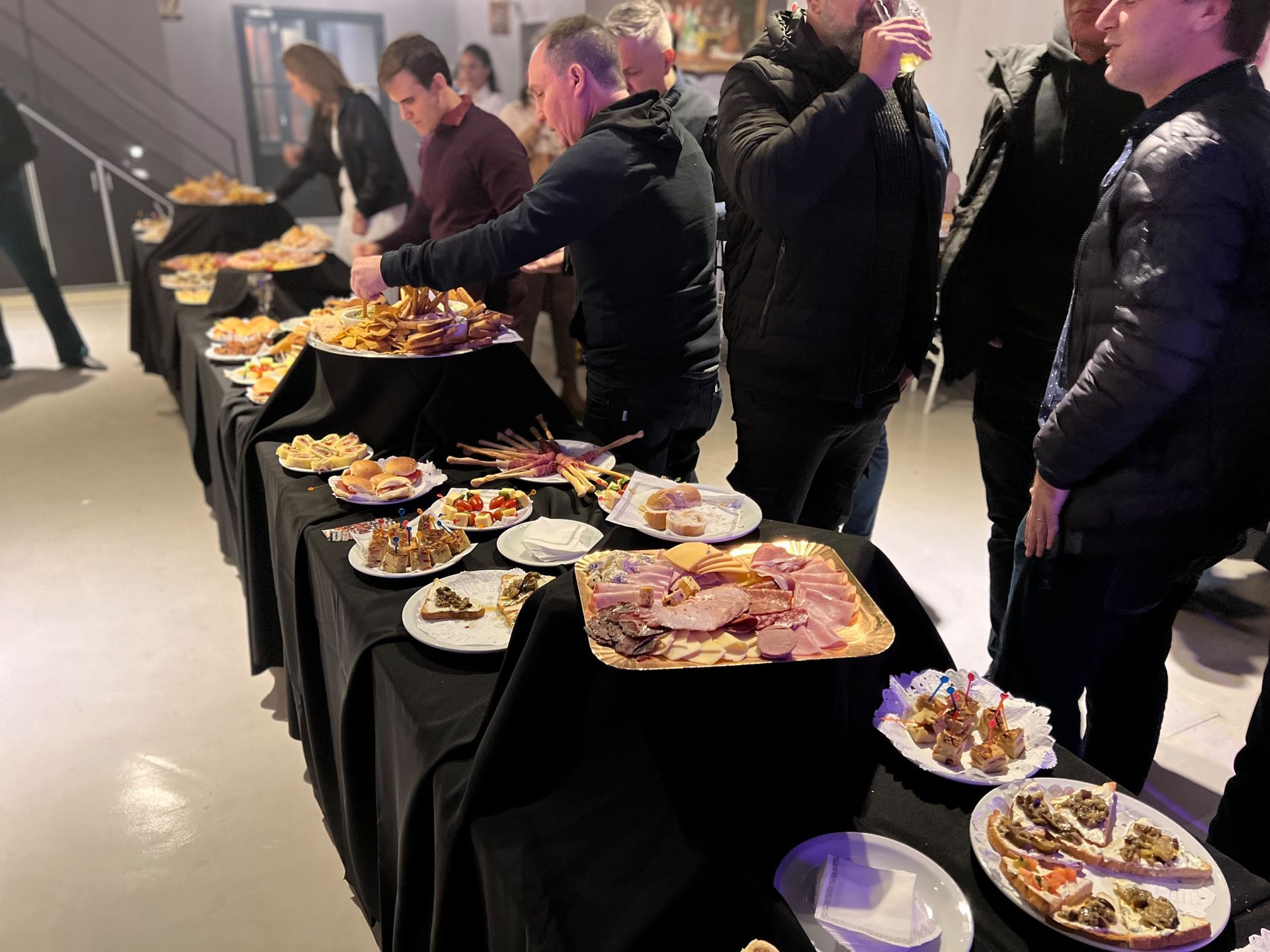 Mesas de bufé repletas de diversos platos mientras los asistentes se sirven en un evento en un espacio cerrado.