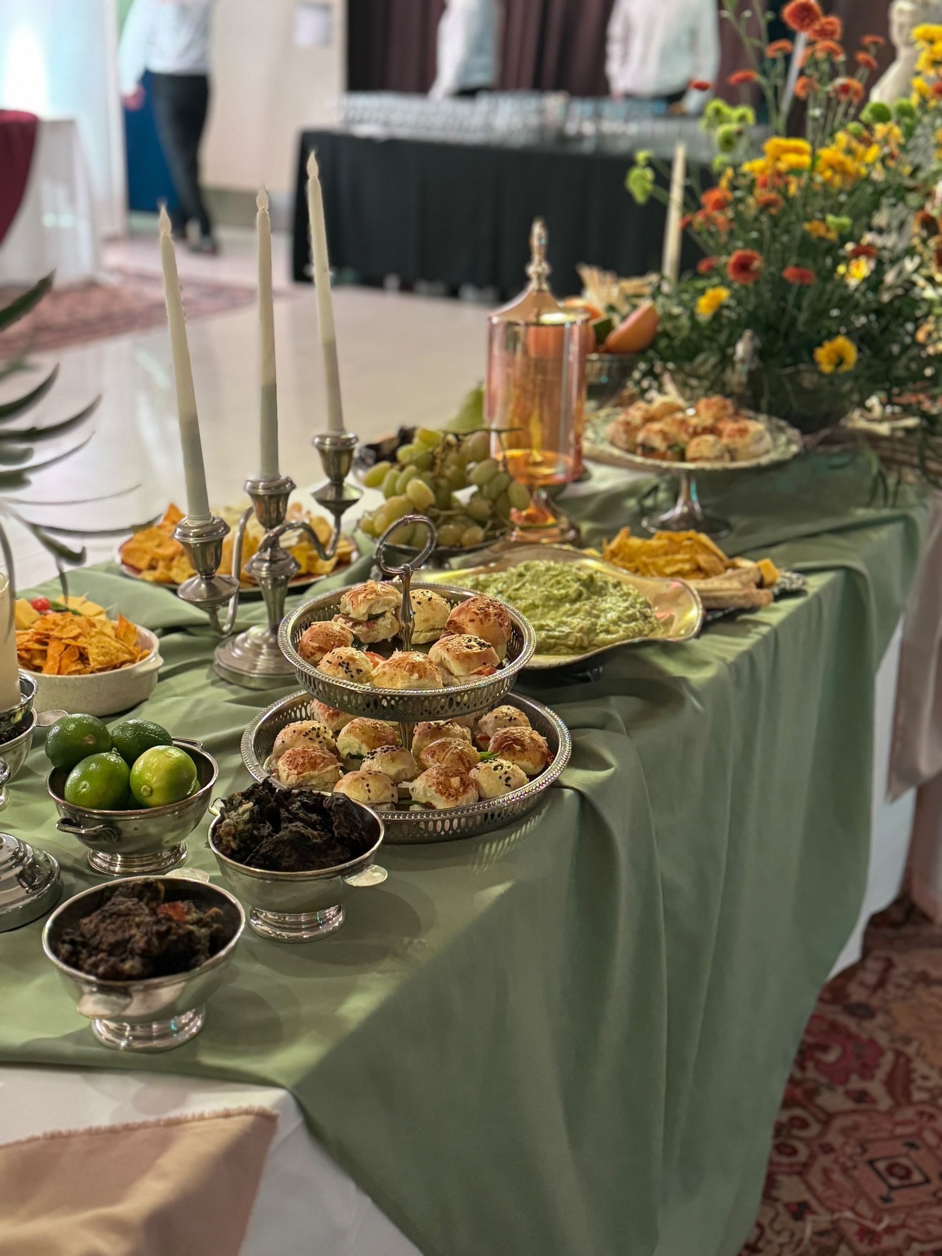 Mesa de buffet con bandejas de comida, fruta, velas y un arreglo floral, preparada en el interior sobre un mantel verde.