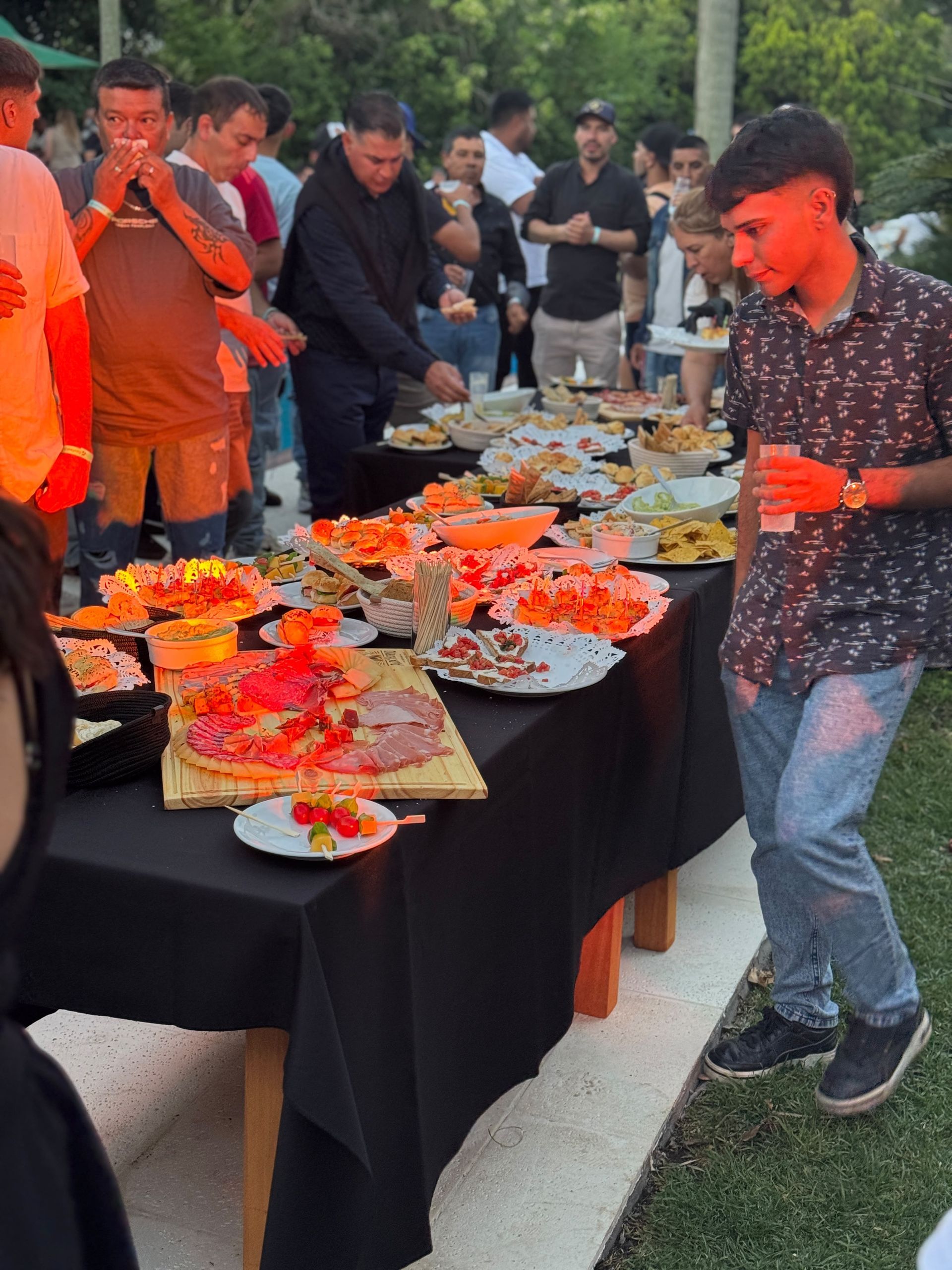 Personas sirviendo y comiendo en un bufé al aire libre, con mesas cubiertas e iluminación naranja