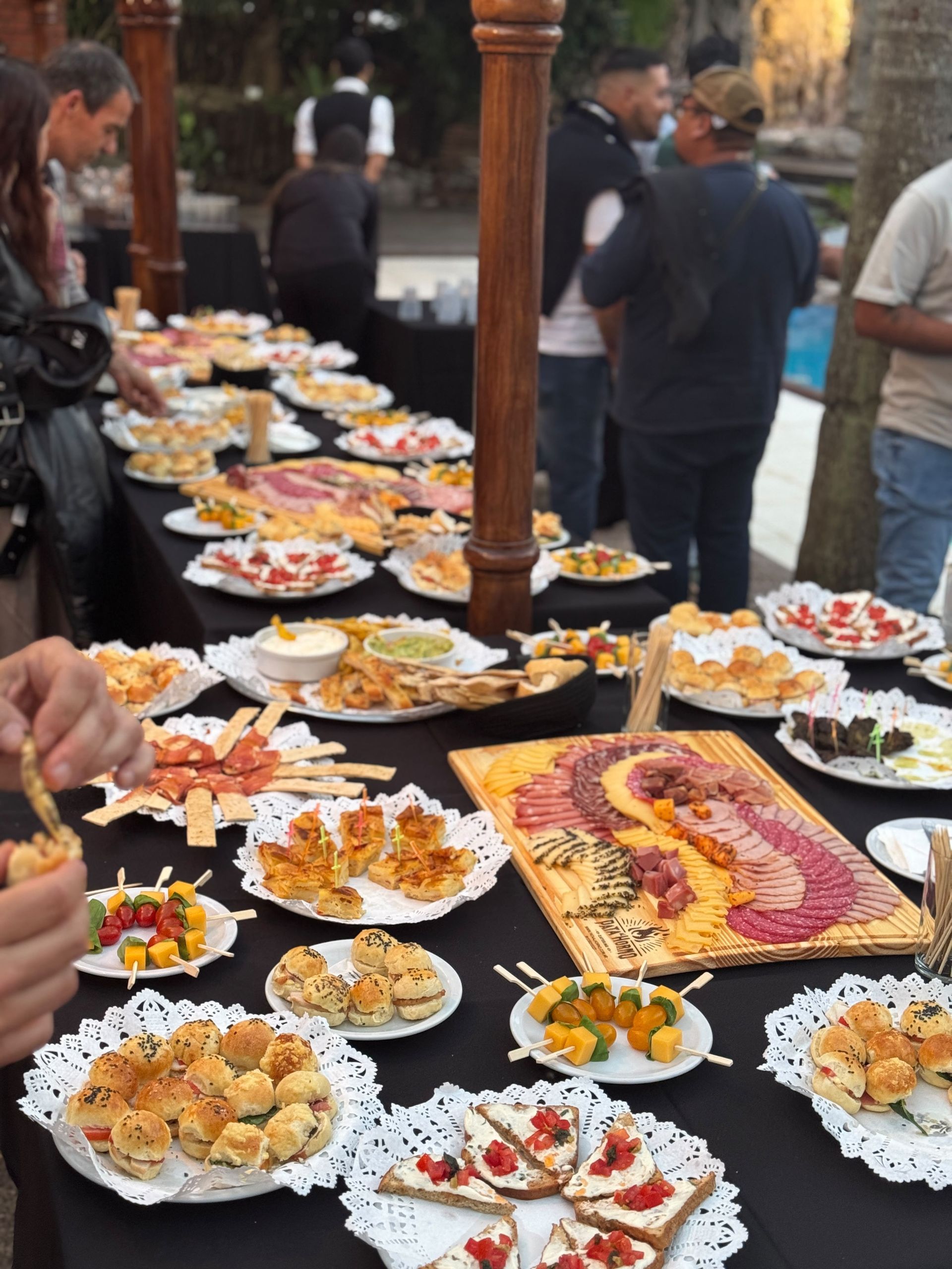 Mesa de bufé al aire libre con muchos platos servidos y fruta, mientras algunas personas están de pie cerca, en un mercado o jardín