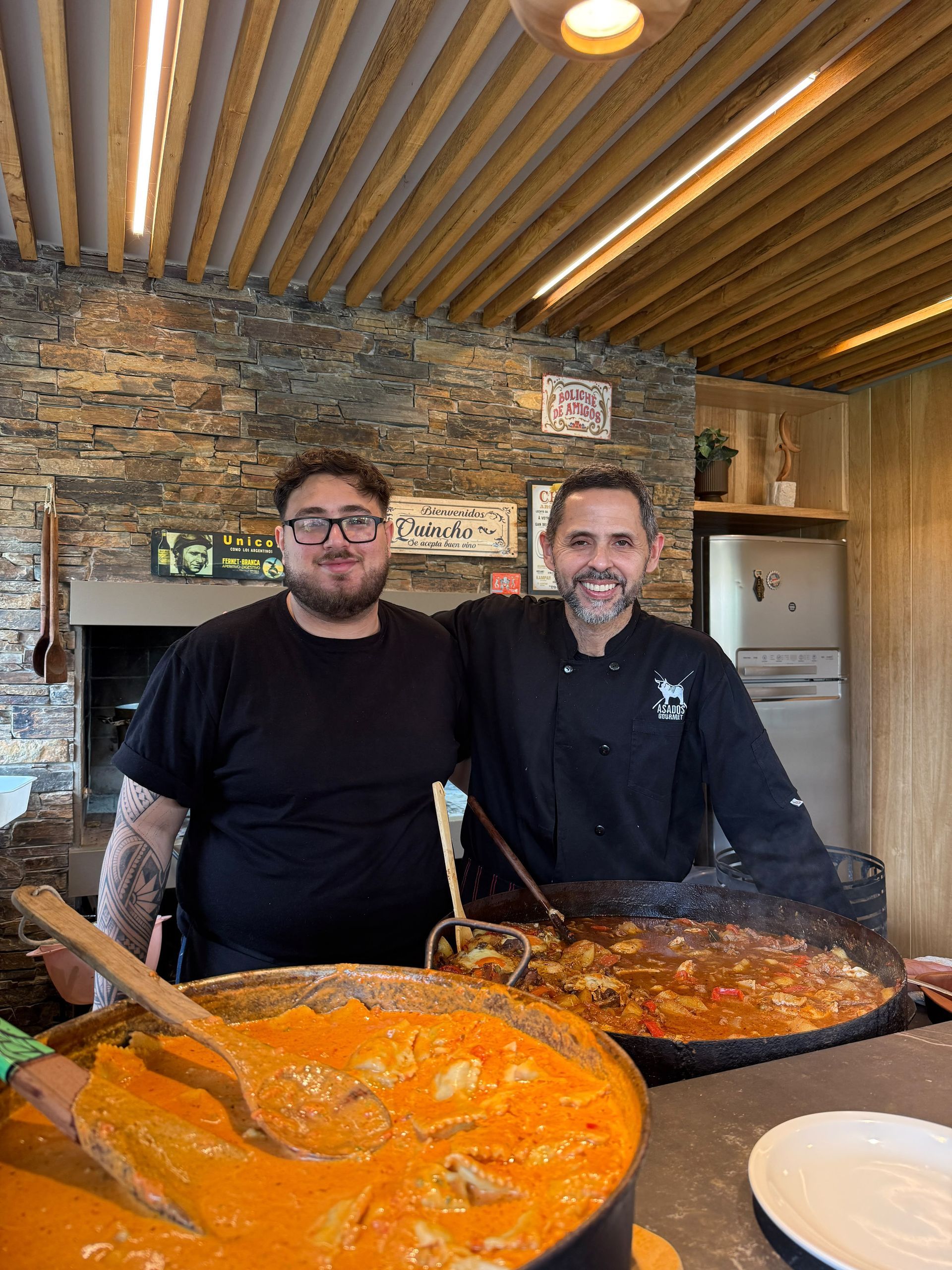 Dos hombres están de pie detrás de unas grandes ollas con guisos de color naranja y oscuro en la cocina de un restaurante rústico.
