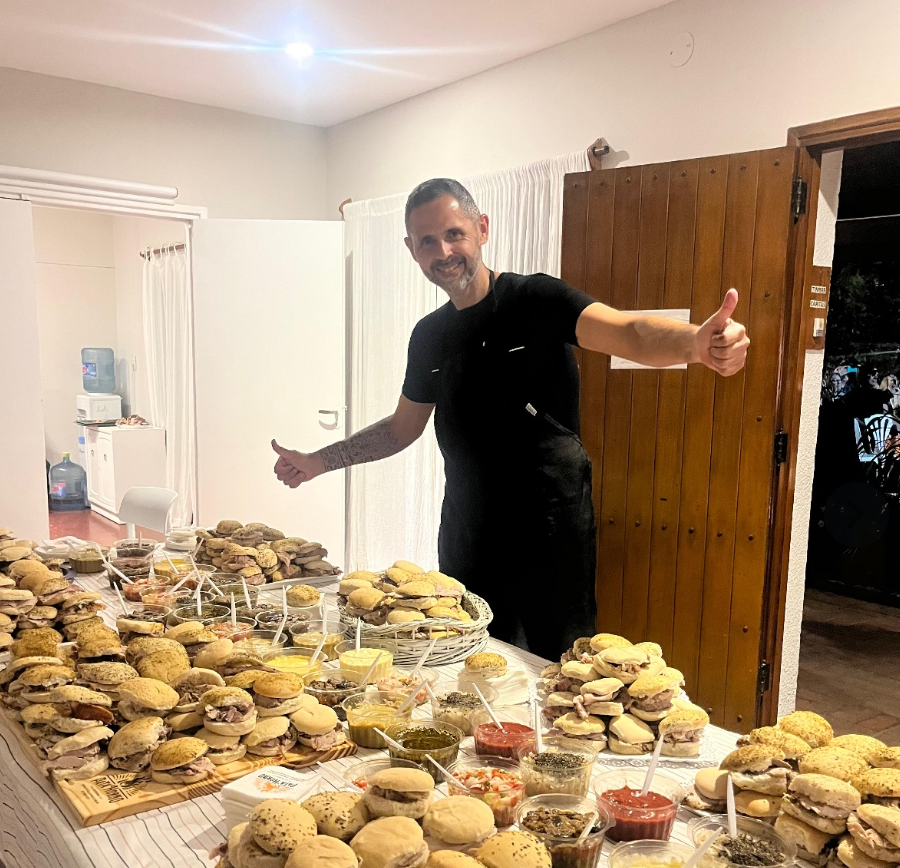 Un hombre de pie junto a unas mesas cubiertas de bandejas con productos horneados en una sala luminosa, levantando el pulgar.