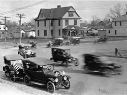 Cars driving at an intersection in a historic town. Buildings and power lines are visible.