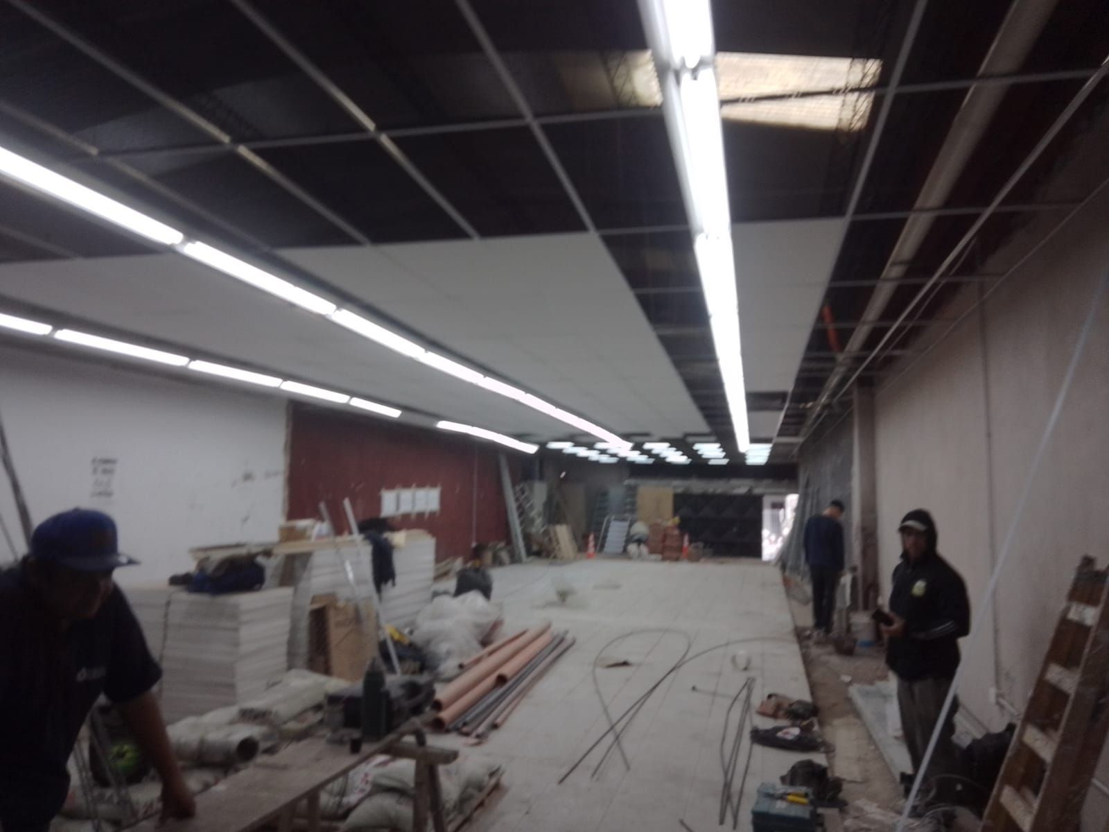 Obreros de la construcción renovando una habitación larga y sin terminar con luces de techo expuestas.