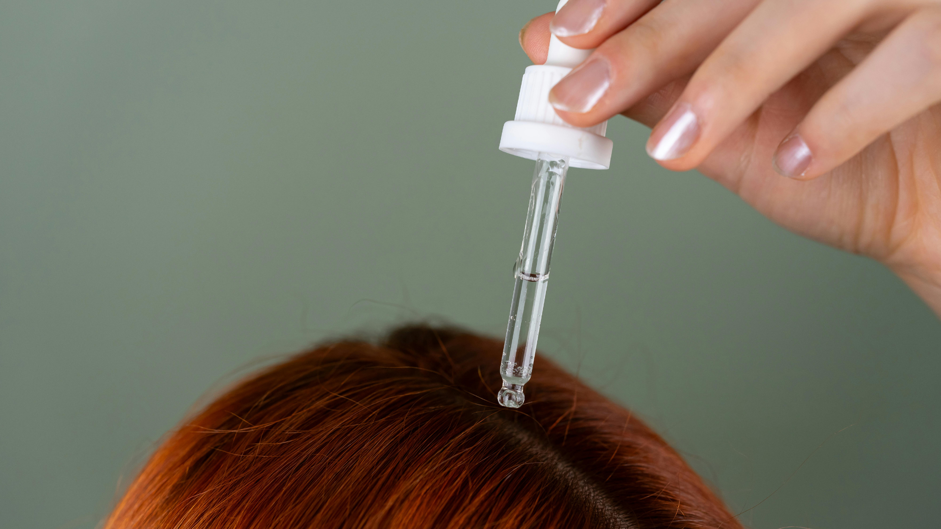 Hand holding a dropper above red hair, applying serum to the scalp
