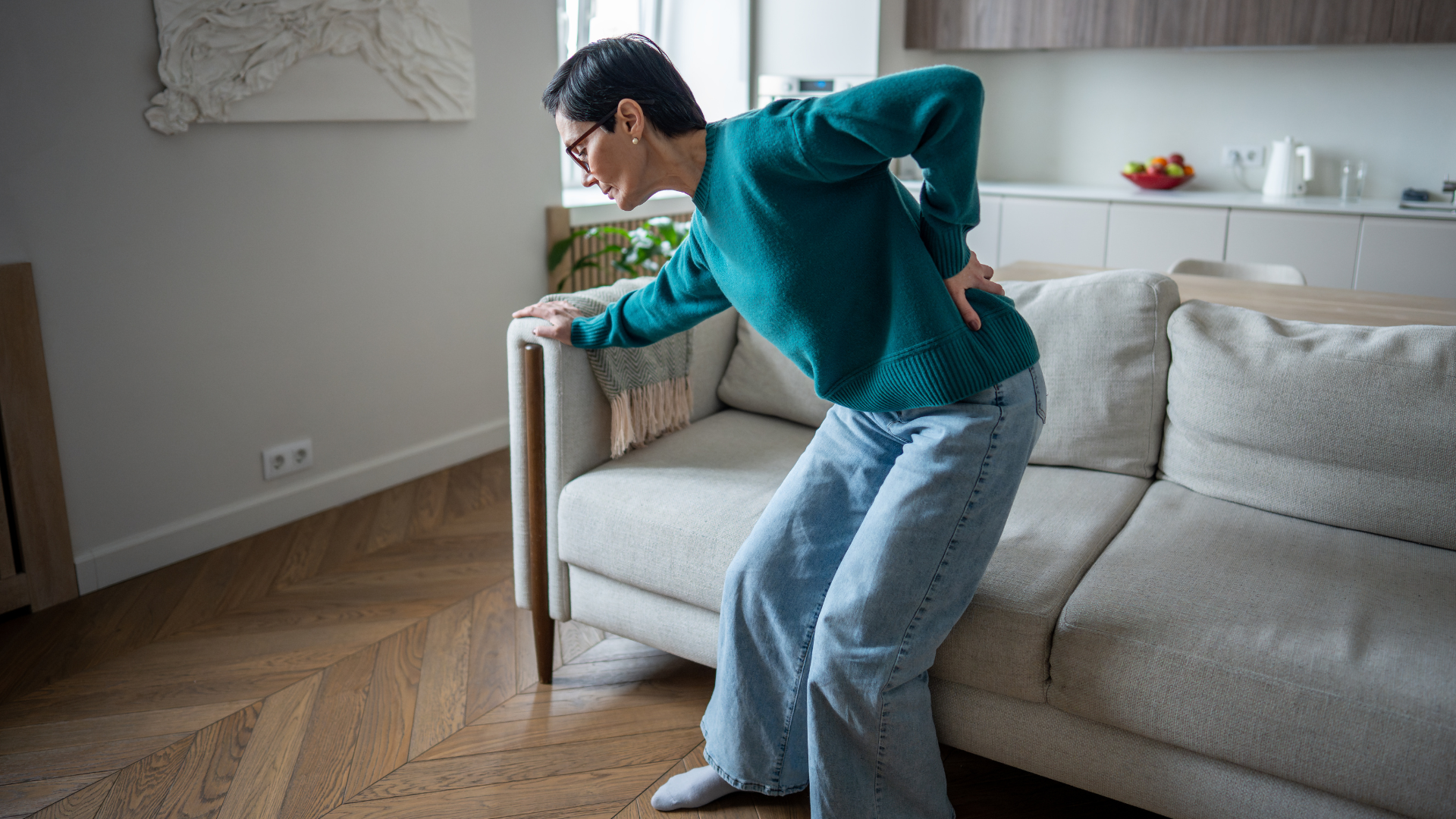 Person holding lower back while standing beside a beige sofa in a bright living room