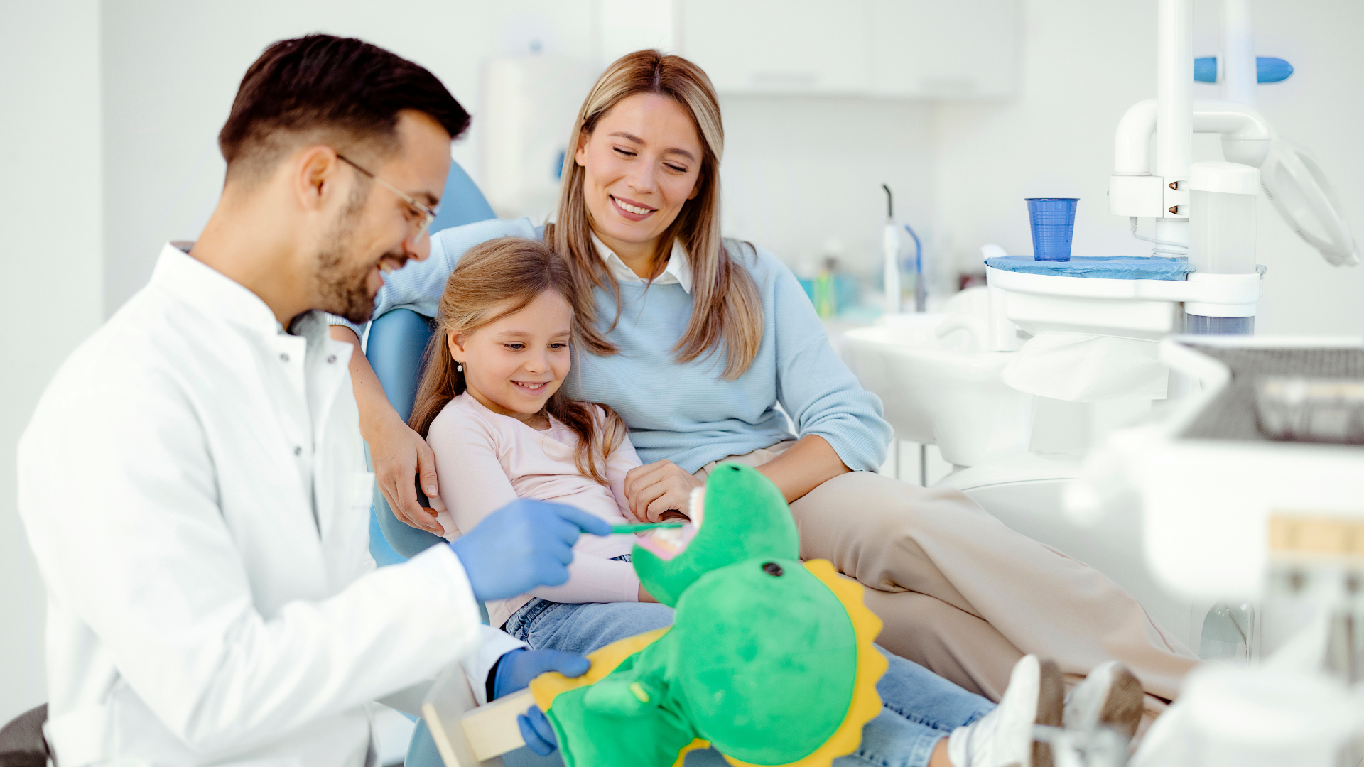 Family visiting a child in a bright hospital room, with a doctor and green stuffed toy nearby